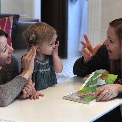 Two adults speak ASL with a hard of hearing toddler girl.
